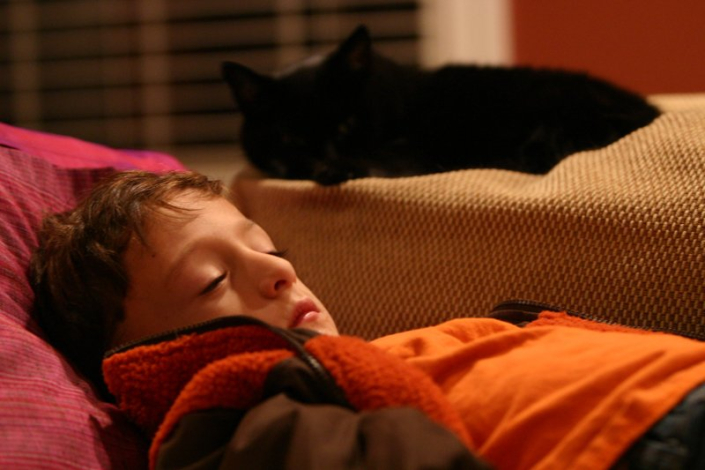 Young boy sleeping on couch with black cat lying behind him